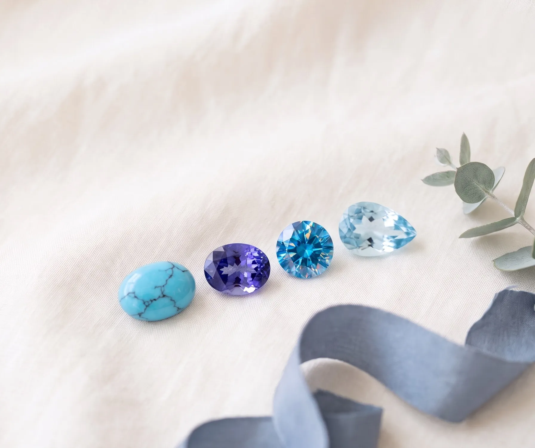 Four loose gemstones in a row on ivory linen, a veined sky-blue turquoise, a violet-blue tanzanite, a bright blue zircon, and a pale blue topaz